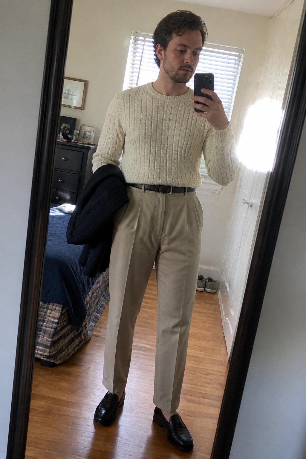 Man in cream cable-knit sweater and beige trousers taking bedroom mirror selfie while holding jacket