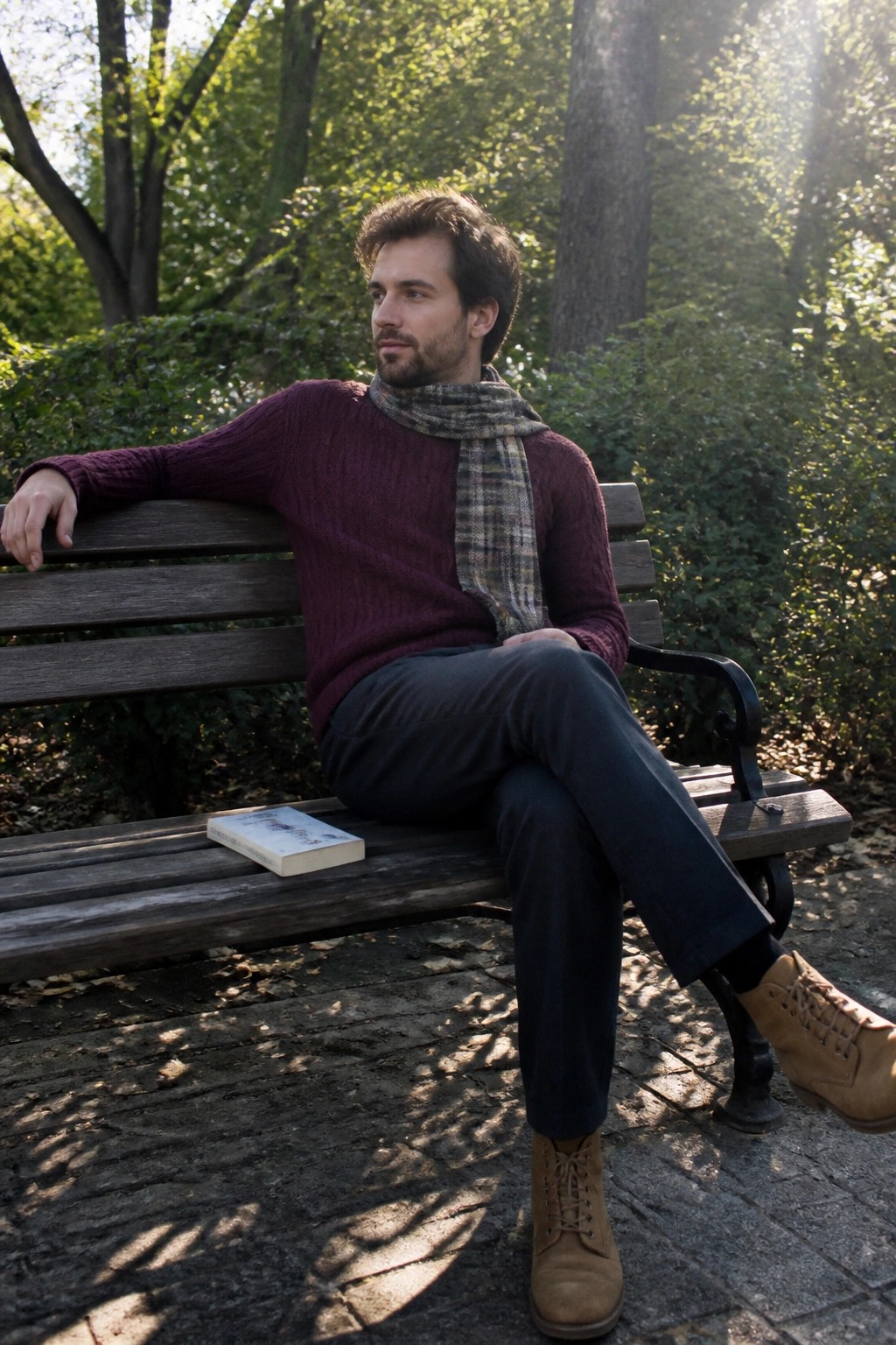 Man in maroon cable knit sweater with plaid scarf tucked in, dark trousers, and tan suede lace up boots seated on wooden bench with book nearby