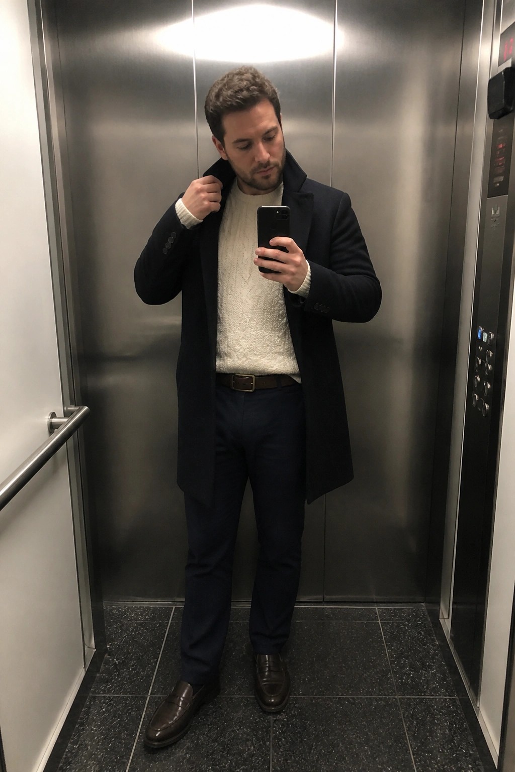 Man in navy overcoat and white sweater taking phone selfie in elevator.