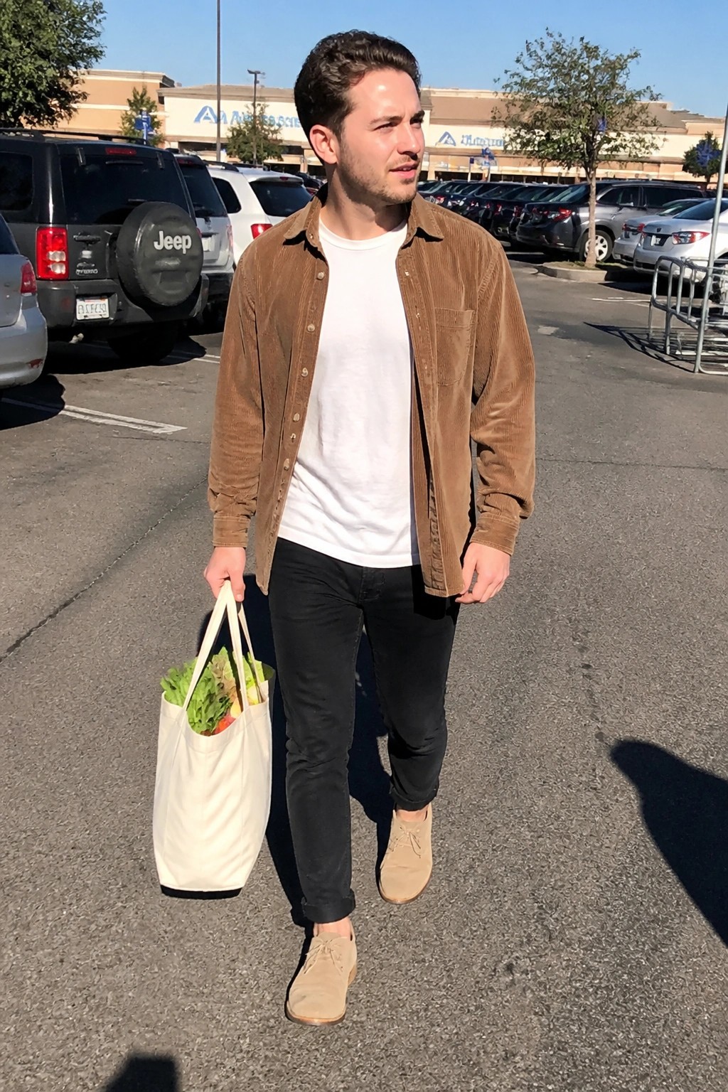 Young man in brown corduroy shirt carrying white tote bag of produce in lot