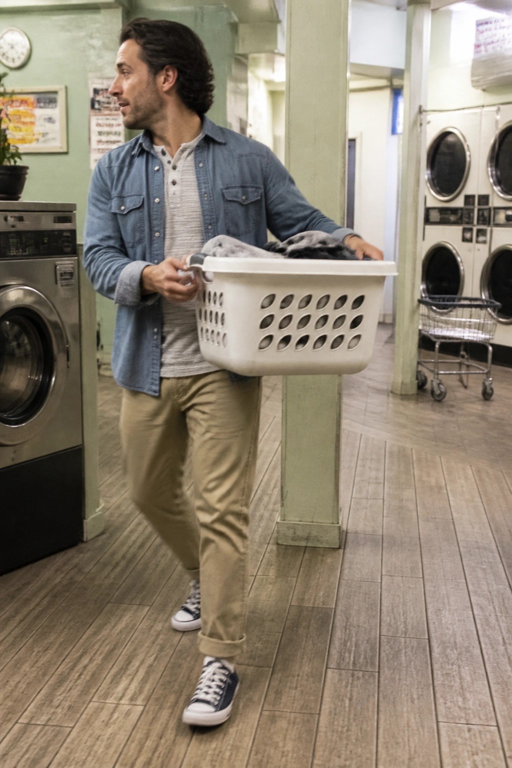 Man wearing open blue denim overshirt over gray striped henley shirt with khaki pants and blue sneakers carrying laundry basket in laundromat setting