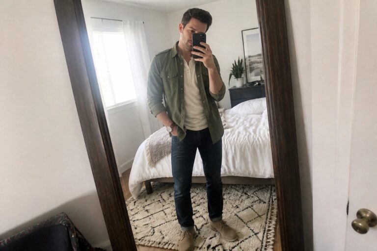 Man in green overshirt over white henley, dark jeans, and boots taking mirror selfie.