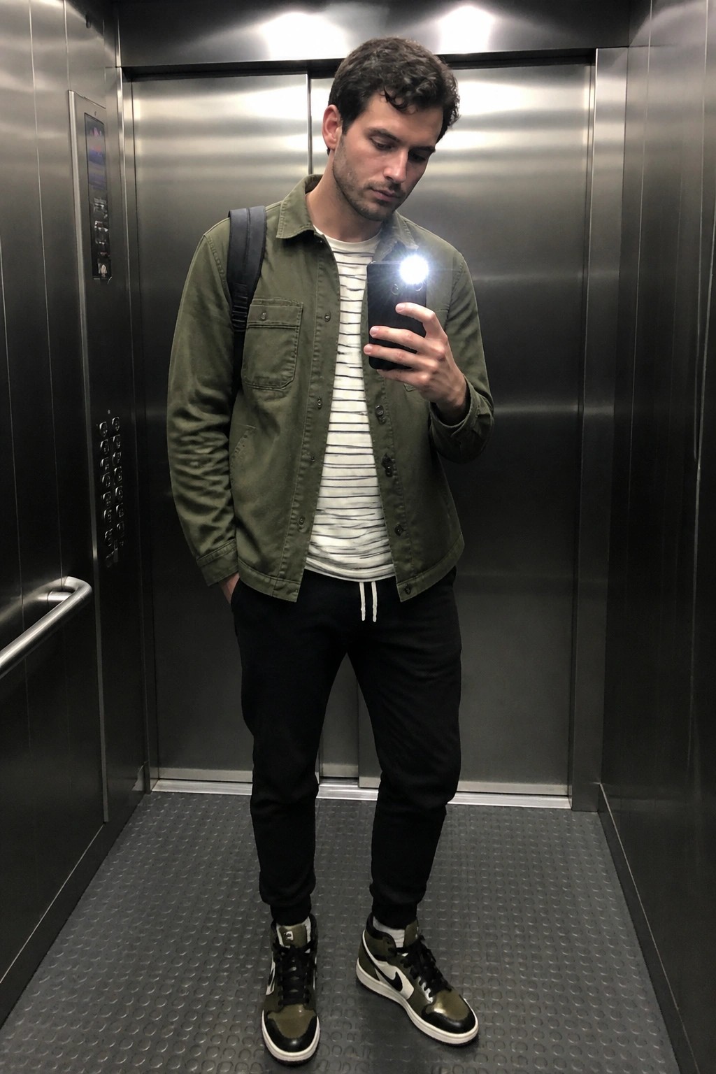 Man in green jacket taking flash selfie inside stainless steel elevator wearing black pants and sneakers