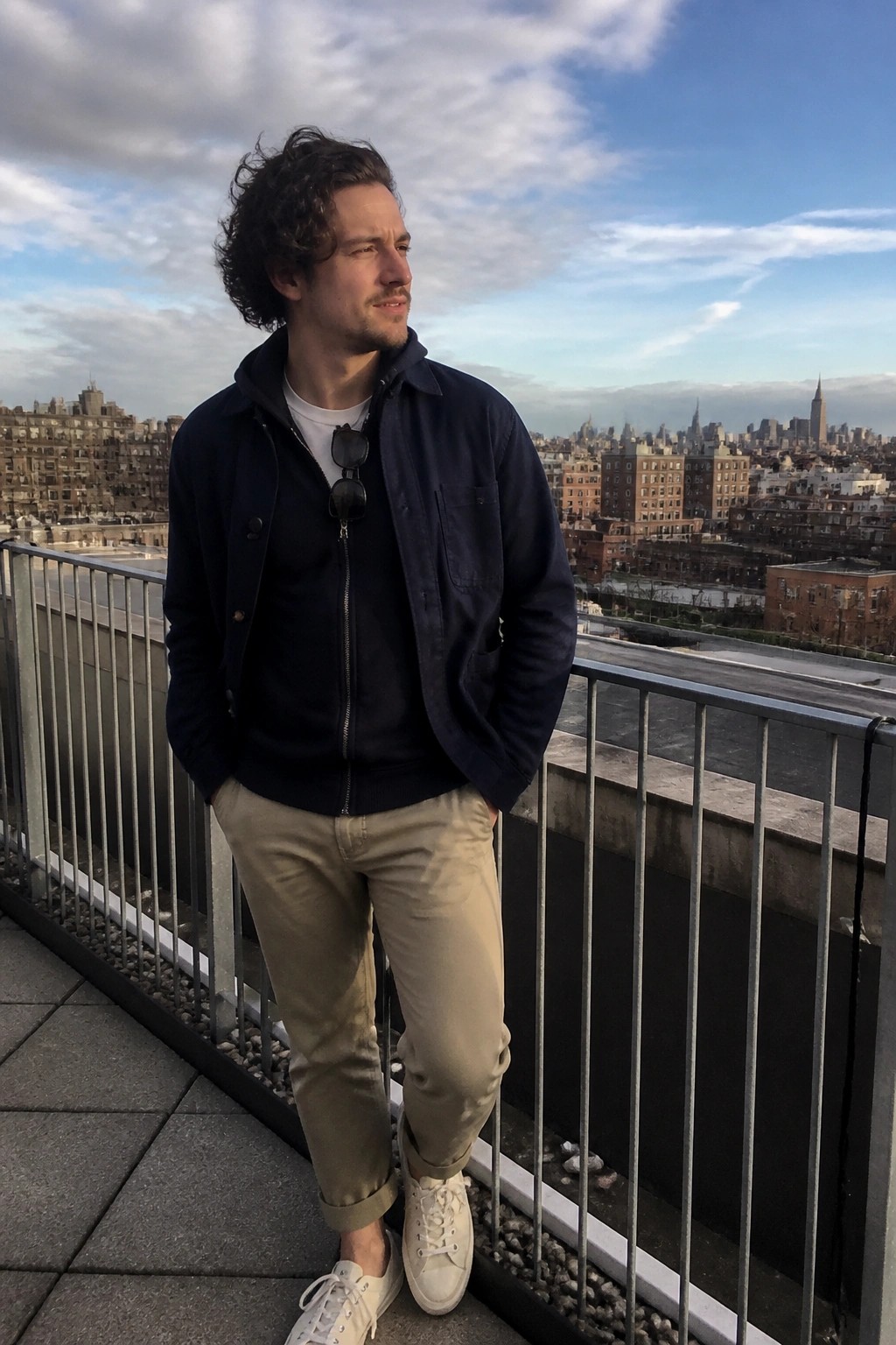 Curly-haired man in navy jacket and beige pants leans on city rooftop railing overlooking skyline