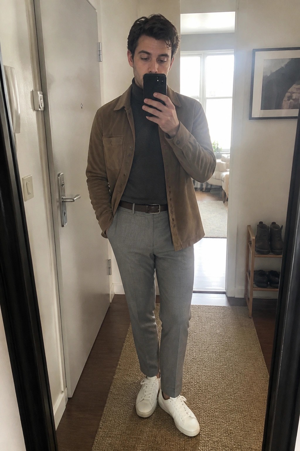 Man in brown jacket and gray pants taking mirror selfie in hallway