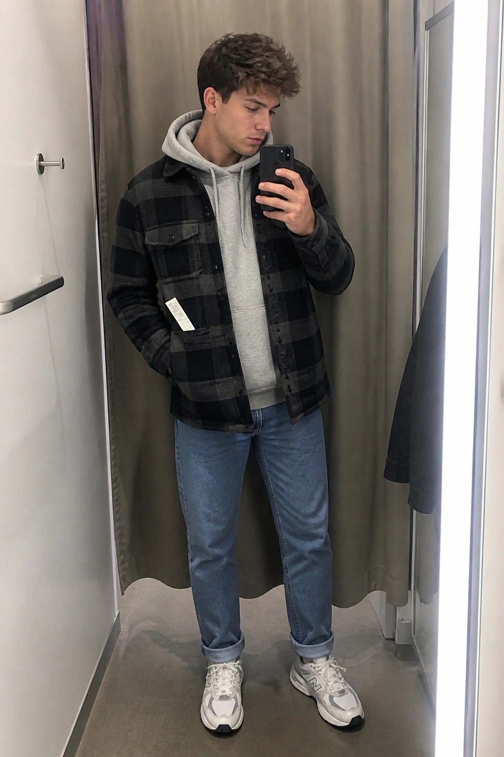 Young man in plaid shirt over gray hoodie taking mirror selfie in jeans and sneakers