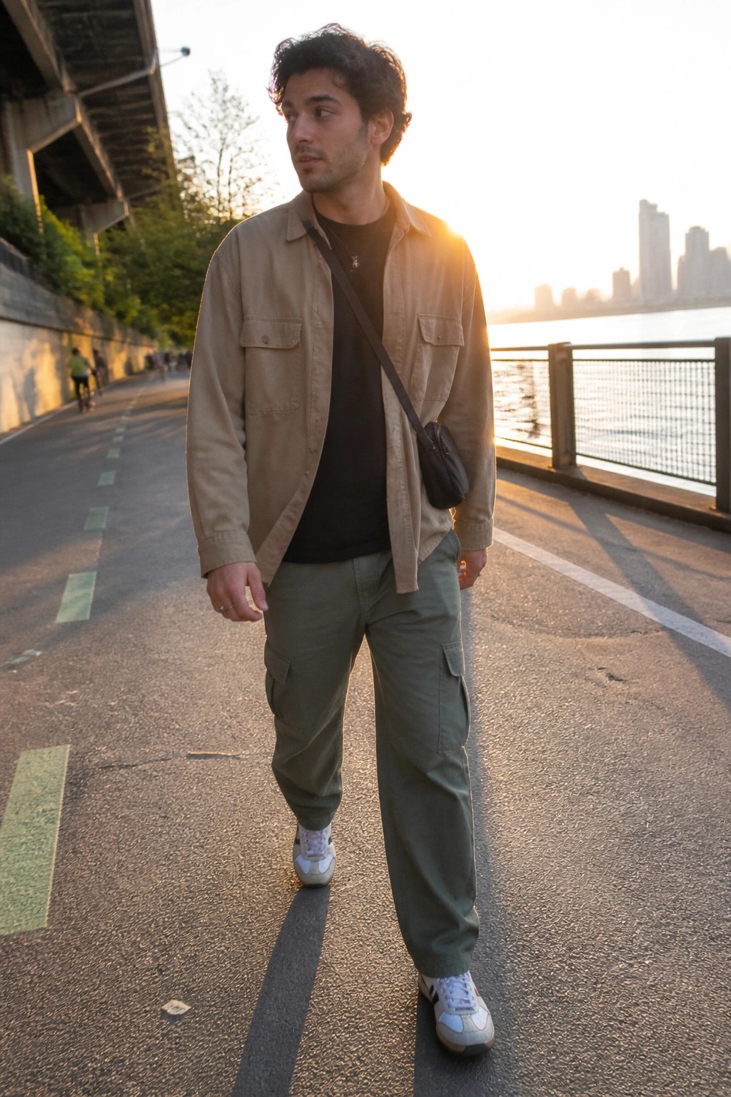 Man in beige overshirt and green cargo pants walking by waterfront at sunset