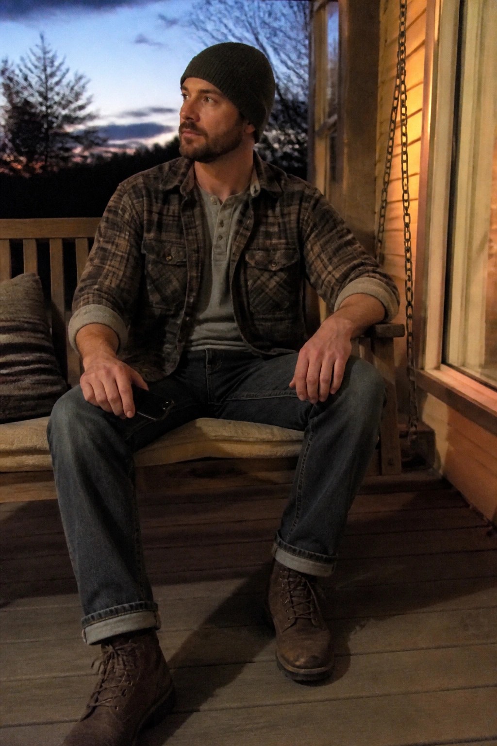 Bearded man in green beanie and plaid shirt sits on porch holding phone at dusk