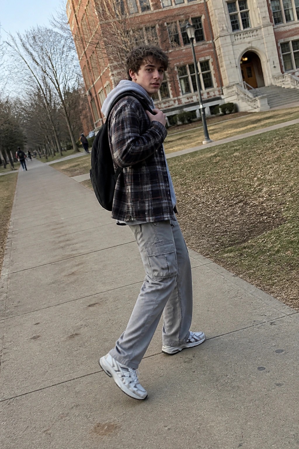 Young man in brown plaid overshirt layered open over gray hoodie paired with gray cargo pants and white sneakers while carrying a black backpack on a paved walkway.