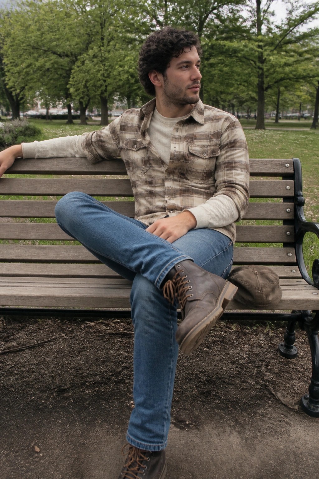 Curly-haired man in plaid shirt and jeans lounges on wooden park bench beside cap.