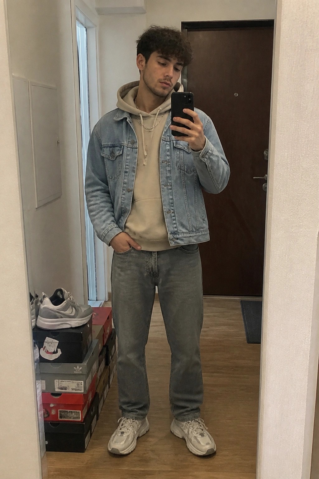 Man wearing light wash denim jacket over beige hoodie with gray jeans white sneakers and silver chain taking mirror selfie in hallway.