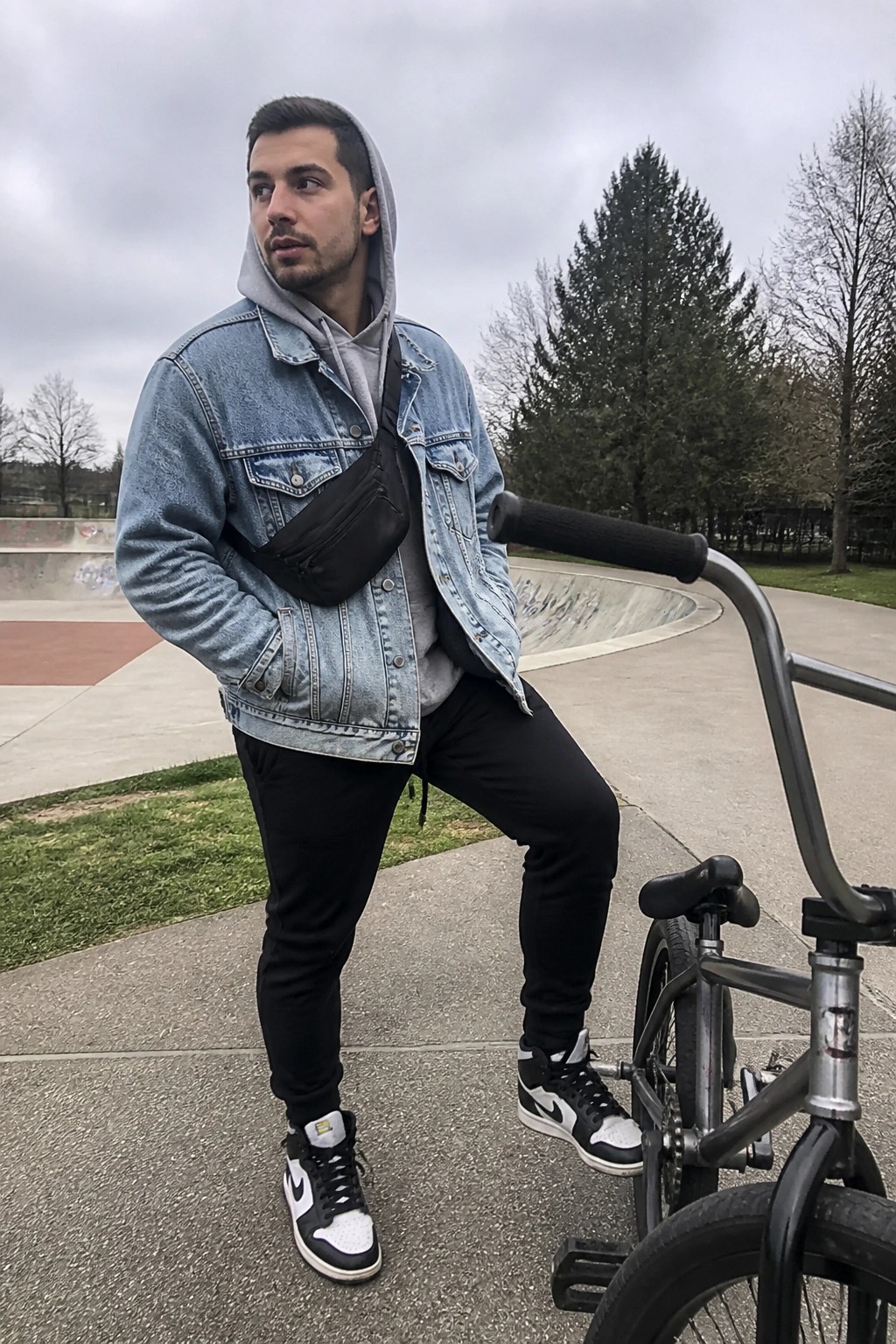 Man in denim jacket and hoodie posing with BMX bike at skatepark