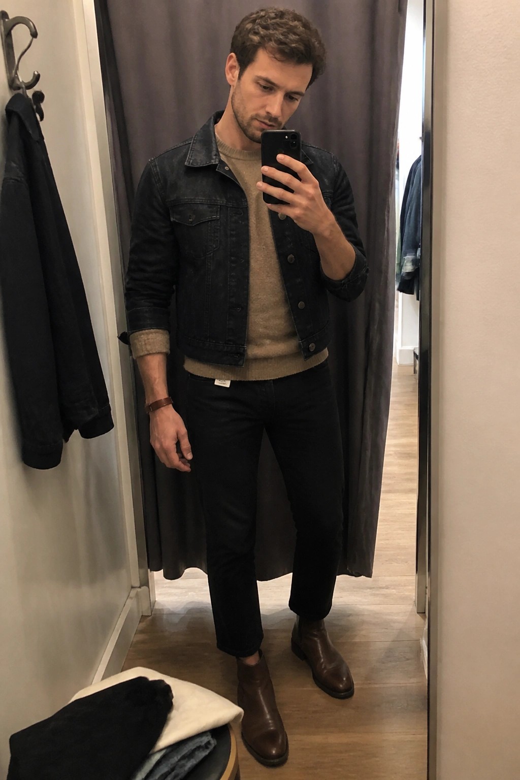 Man in dark denim jacket over beige sweater, black pants, and brown leather boots taking a mirror selfie in a fitting room.