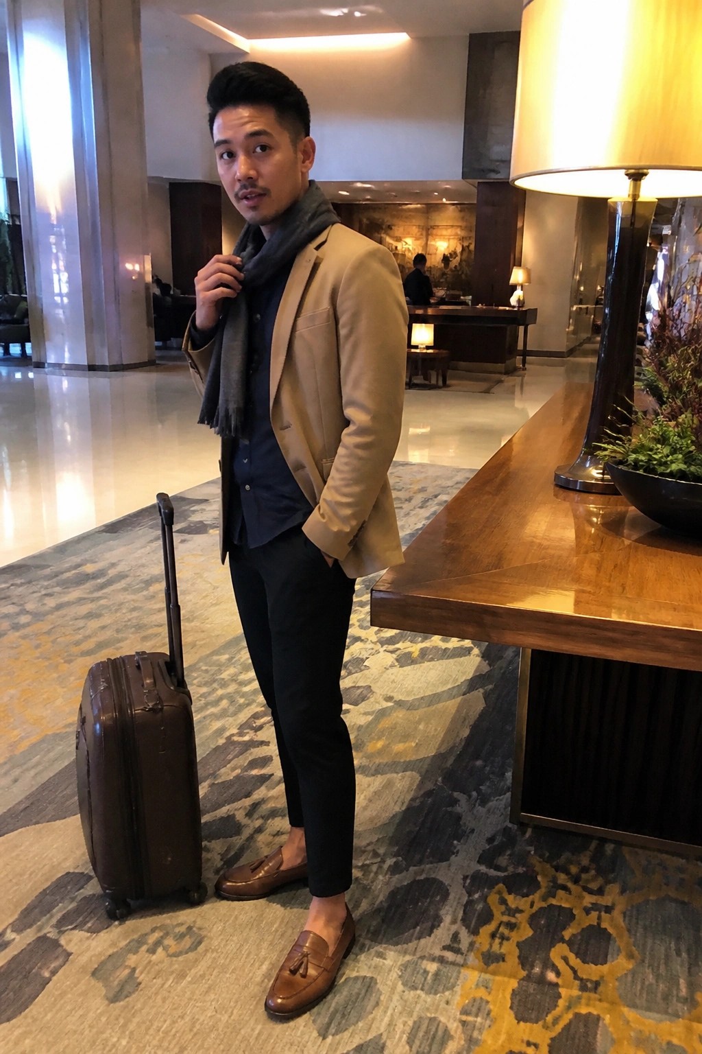 Man in beige blazer and gray scarf with brown suitcase in hotel lobby