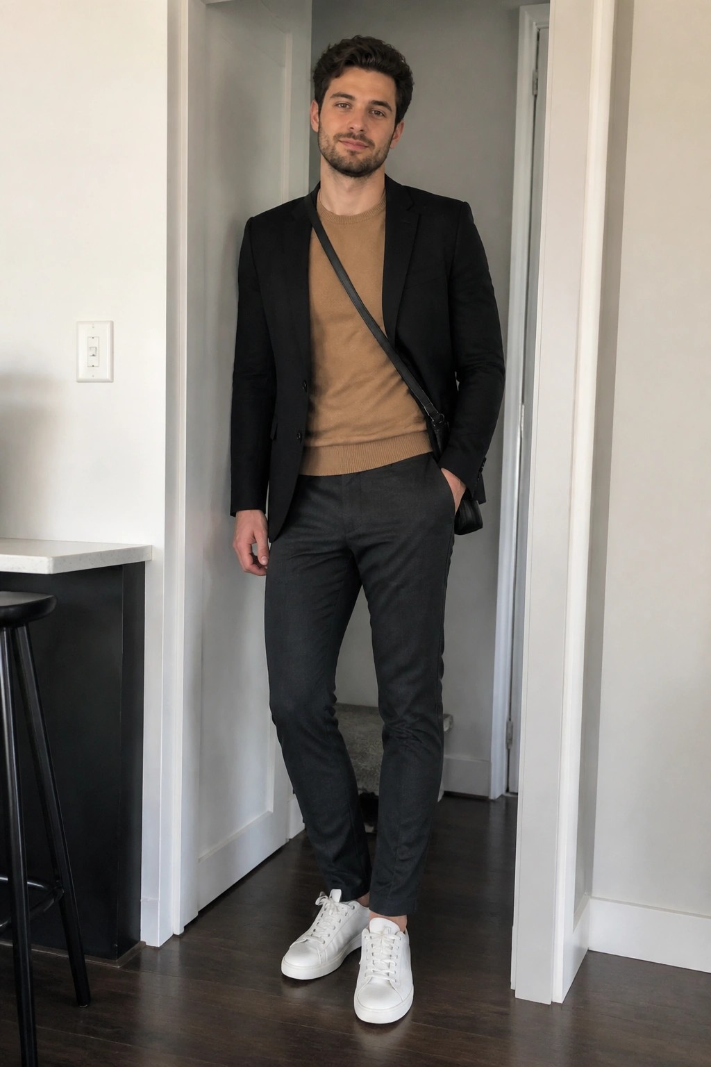 Man in black blazer, tan sweater, gray pants, white sneakers in doorway