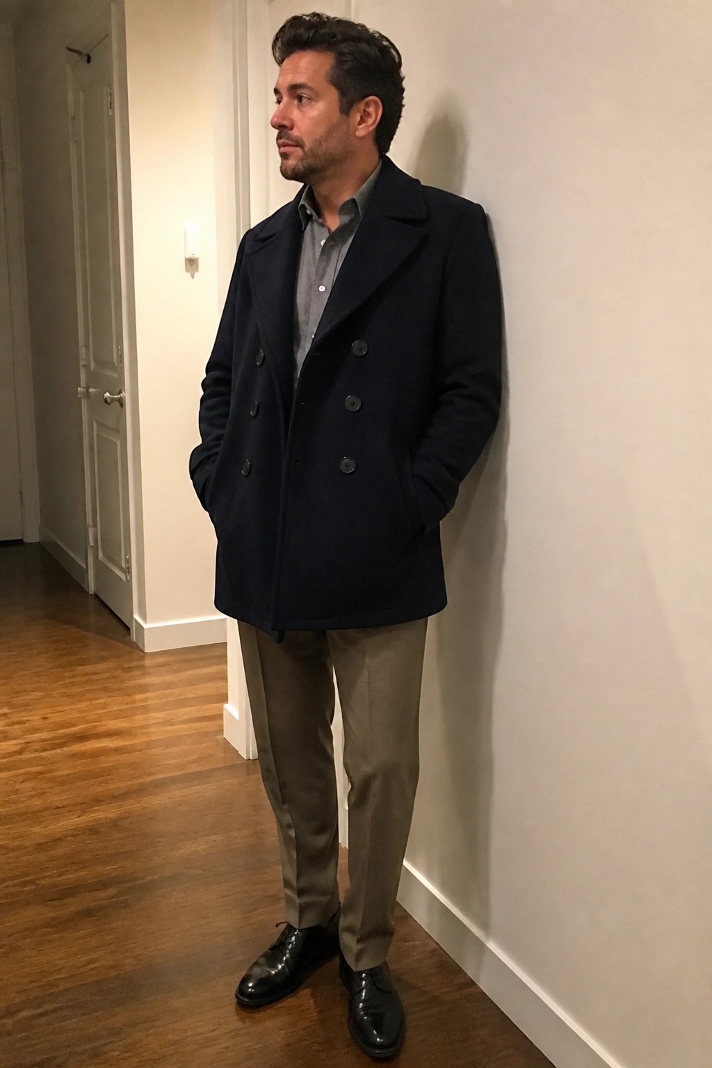 Man in navy peacoat and khaki pants leaning against white wall indoors