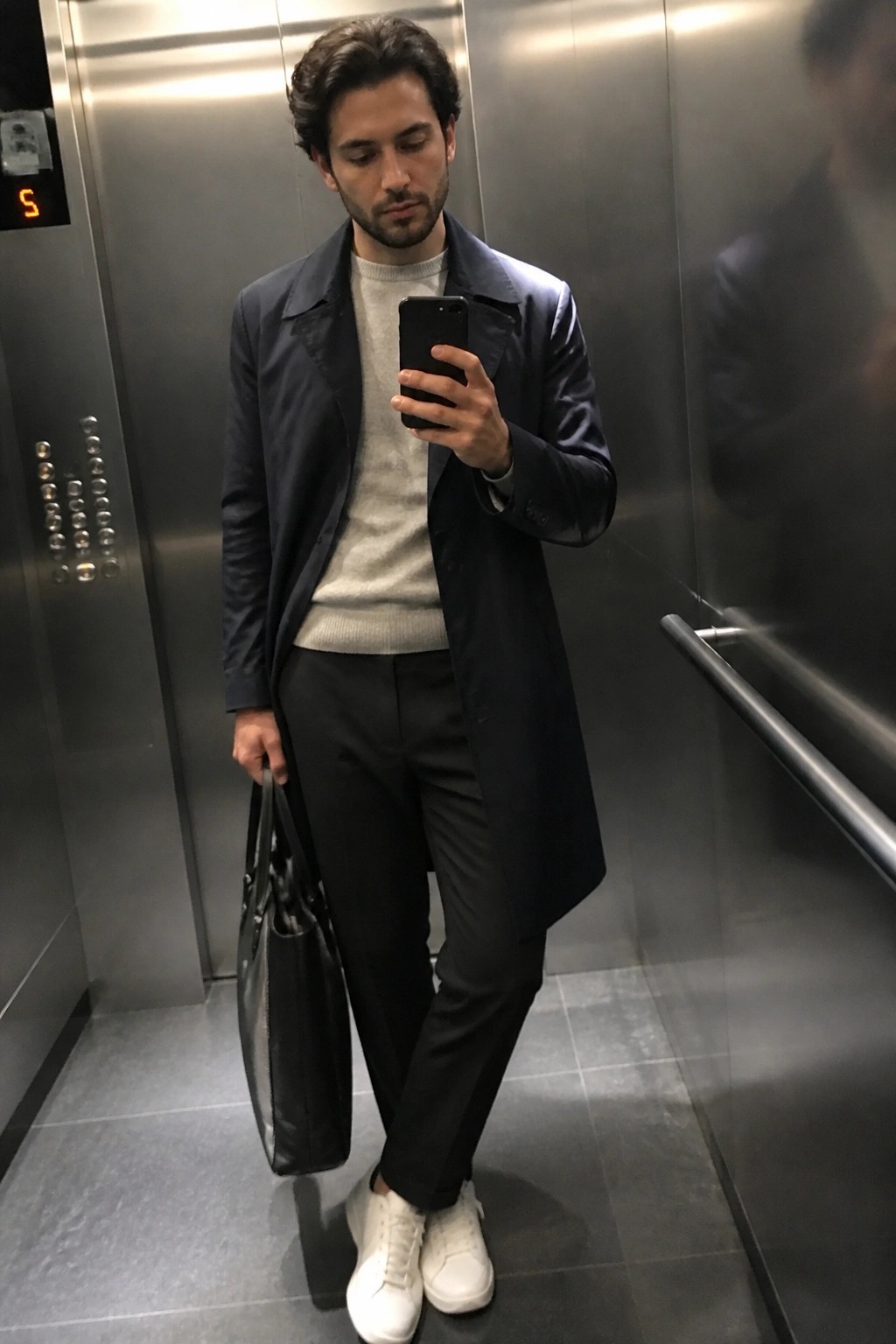 Man in navy overcoat takes mirror selfie in elevator holding black bag.