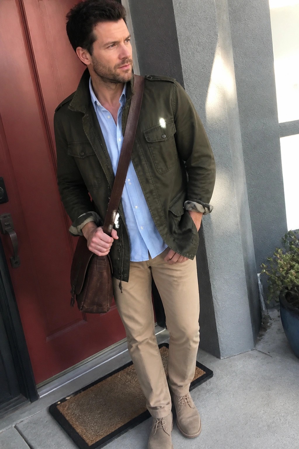 Man in green jacket over blue shirt and khaki pants holding brown leather bag.