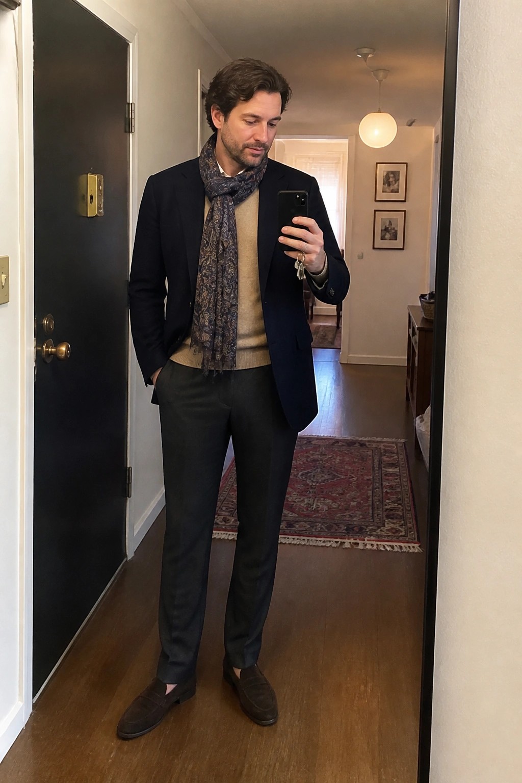 Man in navy blazer and patterned scarf taking mirror selfie in hallway.