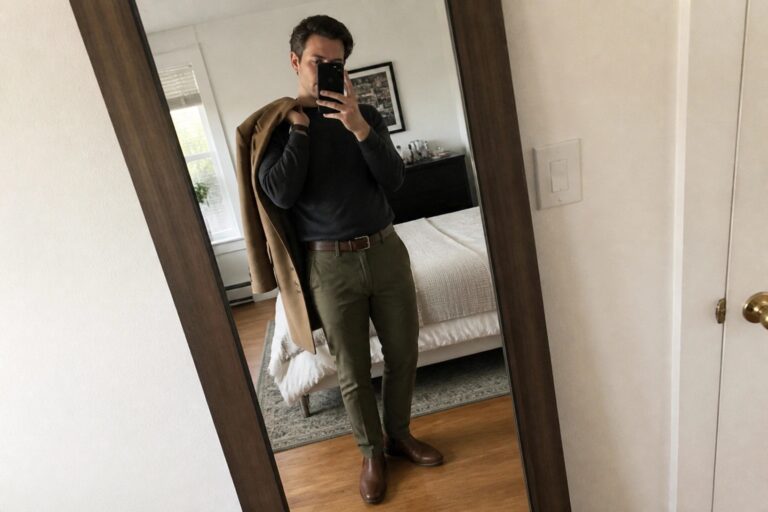 21 Practical Fall Capsule Wardrobe Outfits for Men Built Around Versatile Mix-and-Match Pieces Man in gray sweater and green pants holding tan coat for mirror selfie