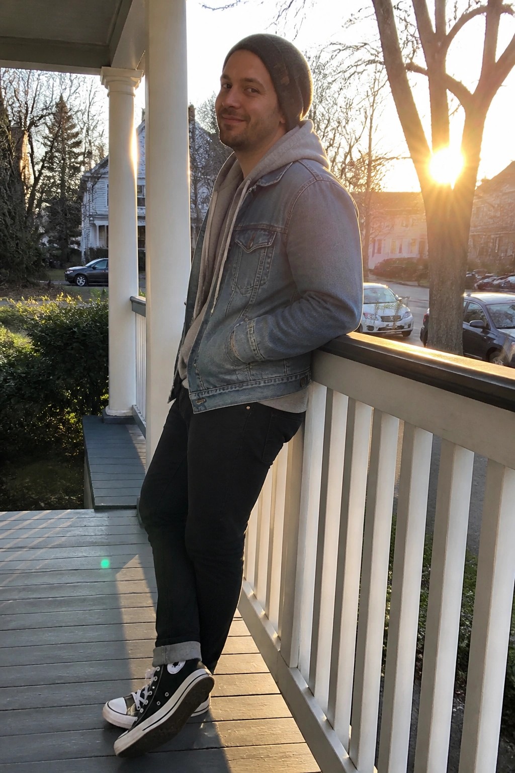 Man in denim jacket and beanie leaning on porch railing at sunset