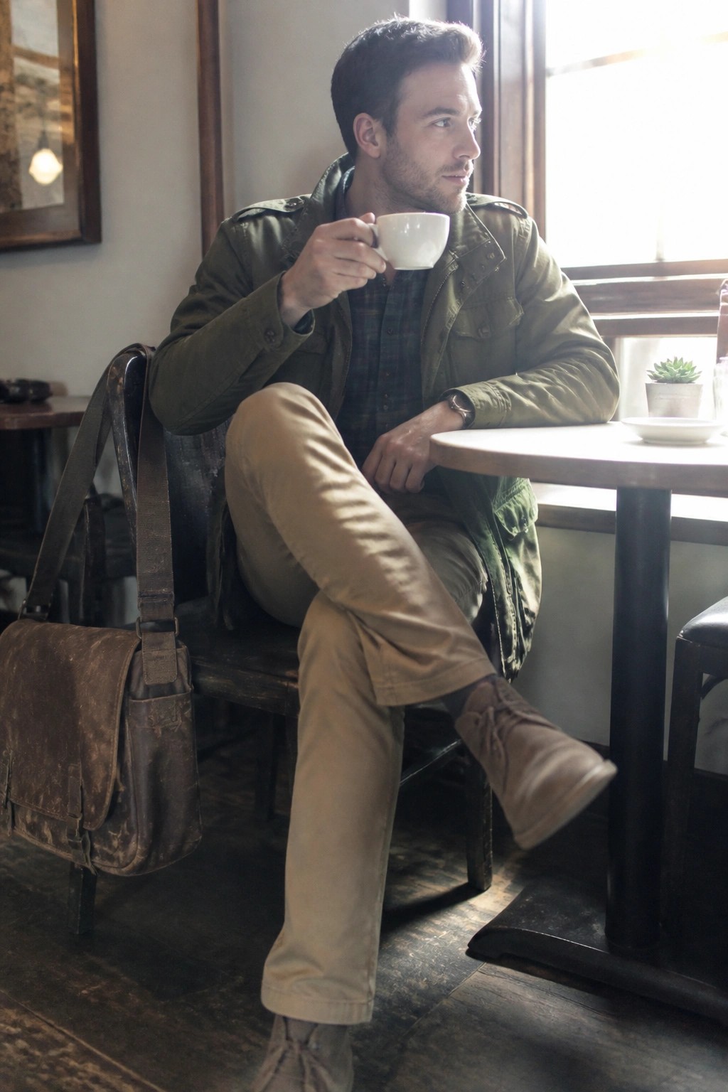 Man in green jacket holds white cup at wooden cafe table with leather bag