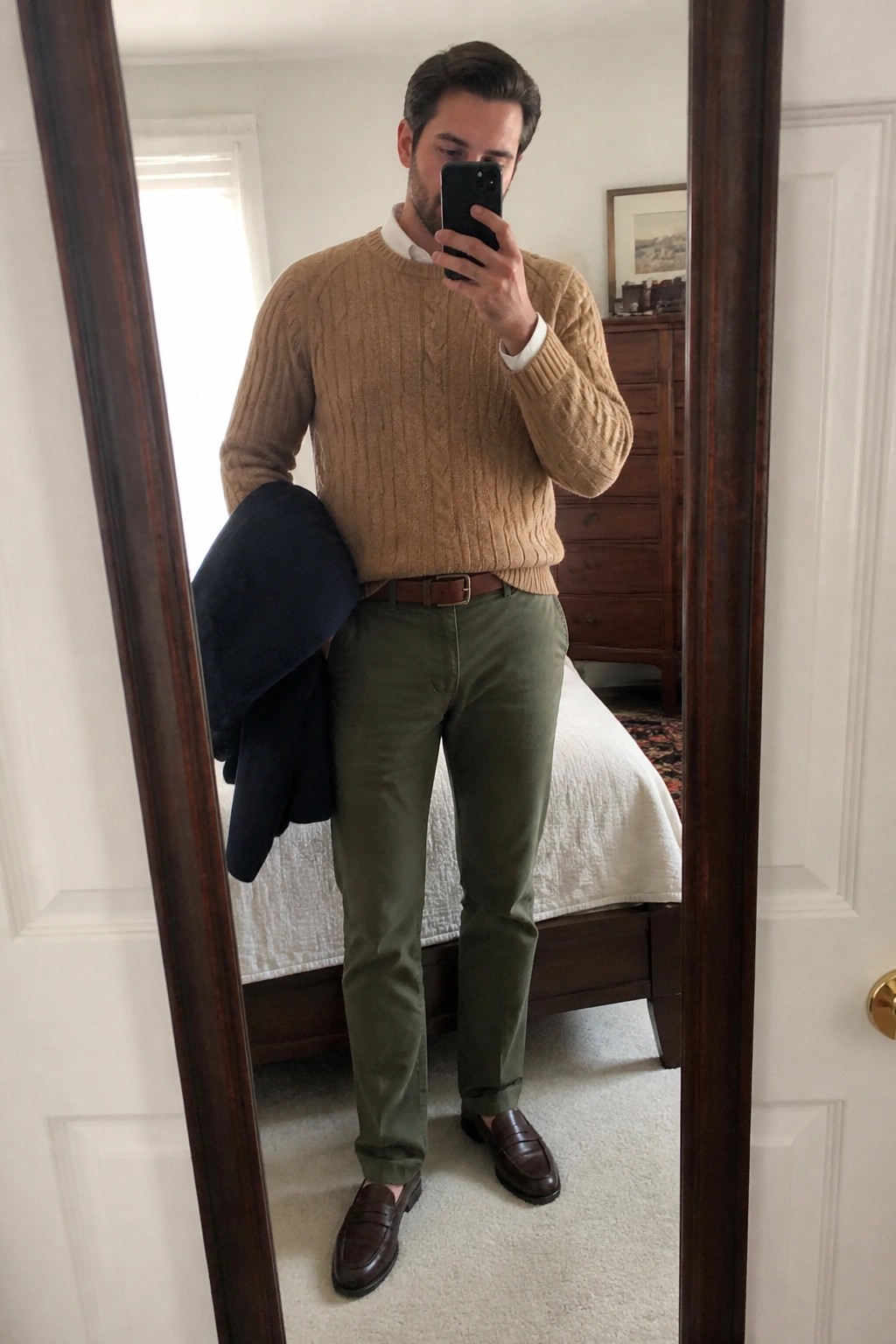 Man in tan cable-knit sweater and olive pants holds navy jacket in mirror selfie.