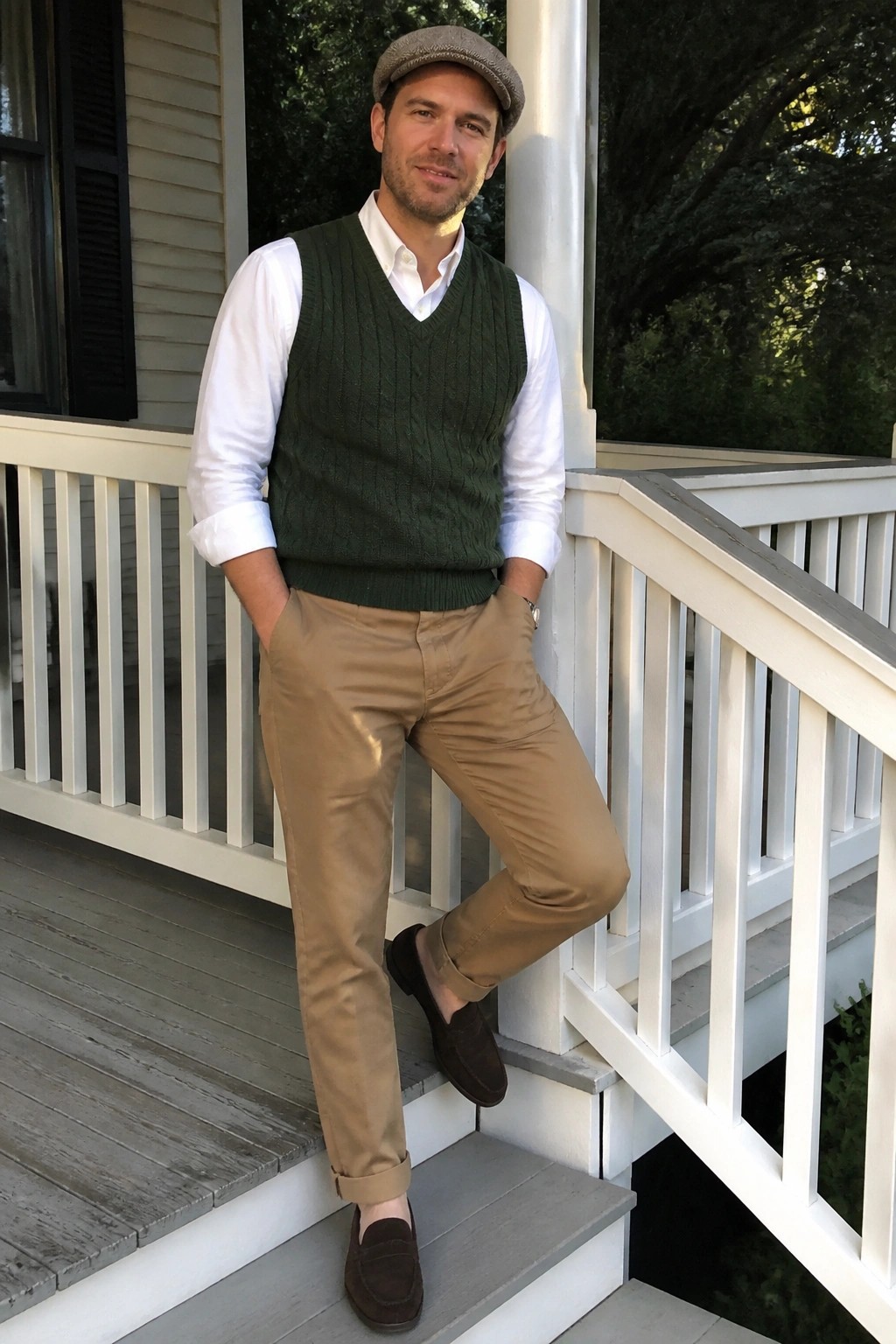 Man in green cable-knit vest, white shirt, tan pants, and flat cap on porch.
