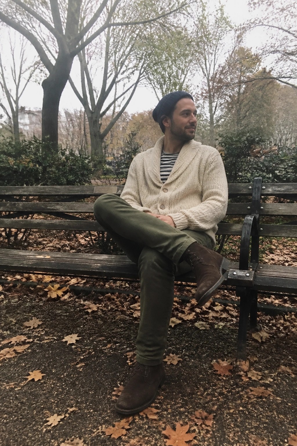 Man in cream knit cardigan and beanie sits on leaf-covered park bench in autumn.