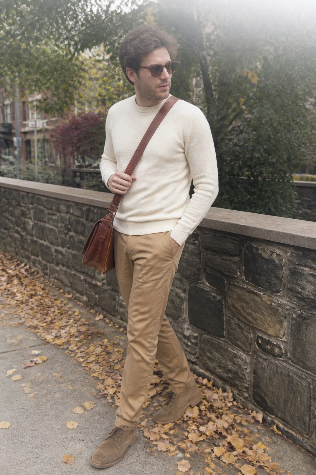 Man in cream sweater and tan pants with brown leather bag on autumn path