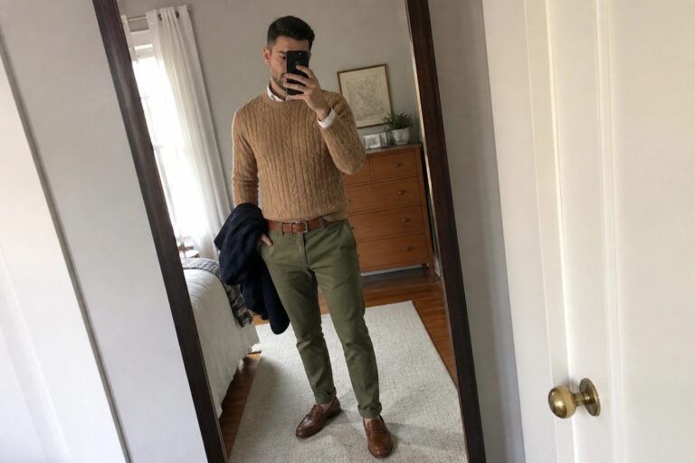 21 Preppy Old Money Fall Outfits for Men Featuring Chinos and Knitwear Man in tan cable-knit sweater and olive pants holds navy jacket in mirror selfie.
