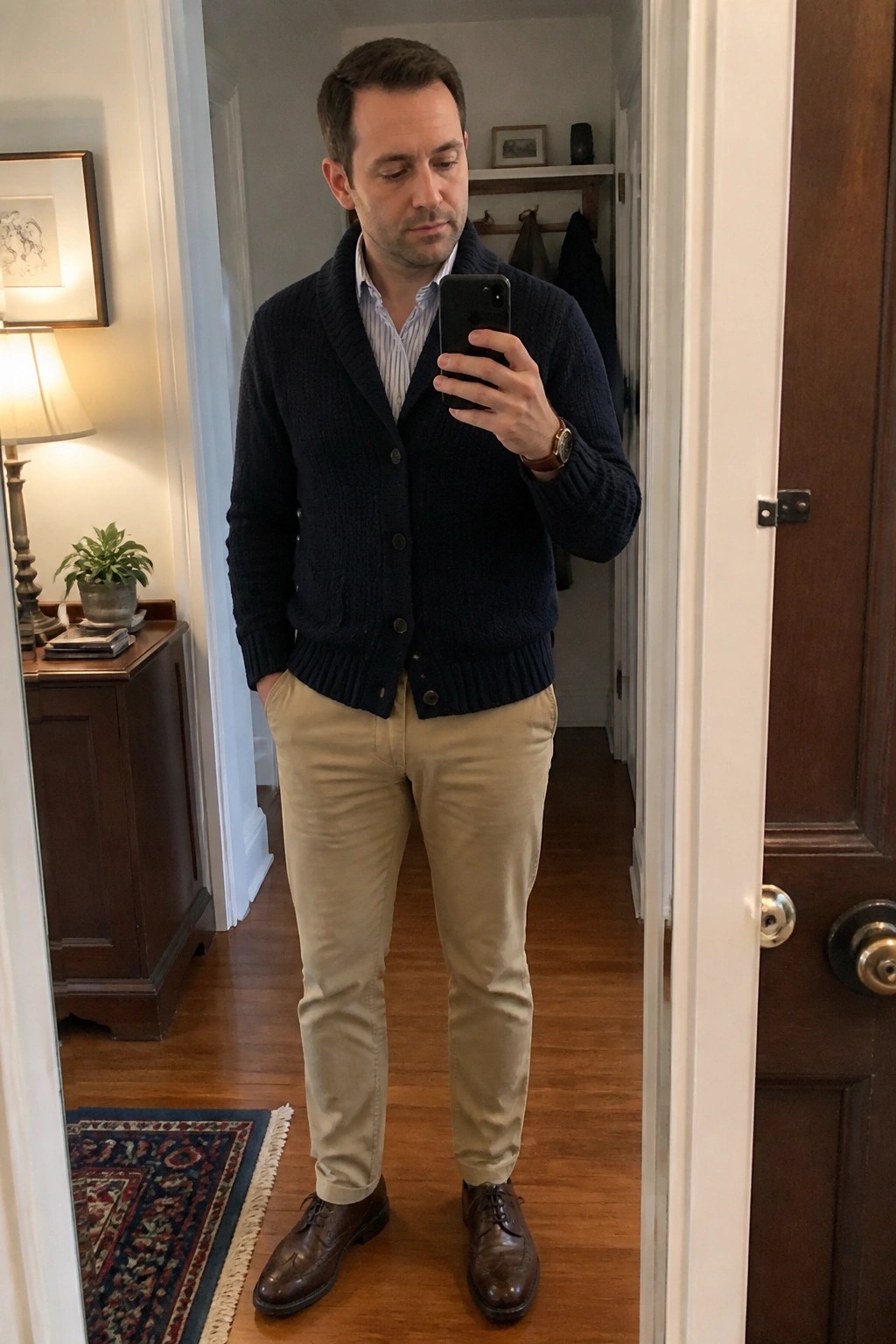 Man in navy cardigan, striped shirt, and khaki pants taking mirror selfie