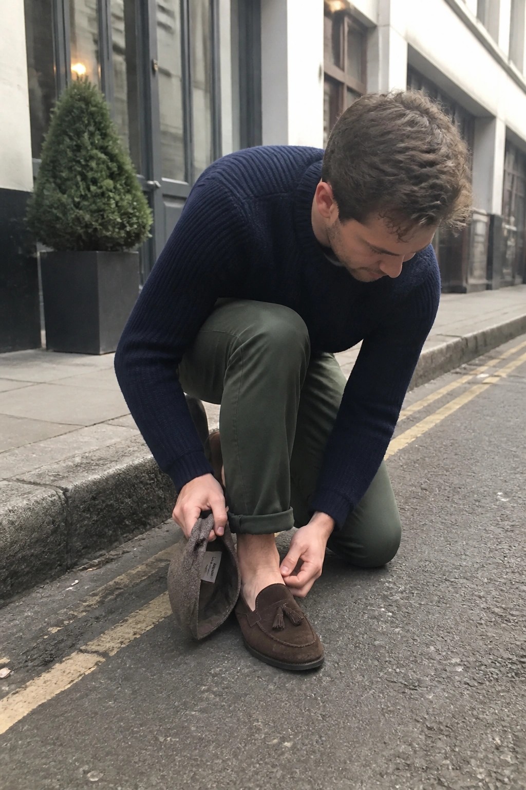 Man in navy sweater and green pants adjusting brown suede tassel loafers on sidewalk
