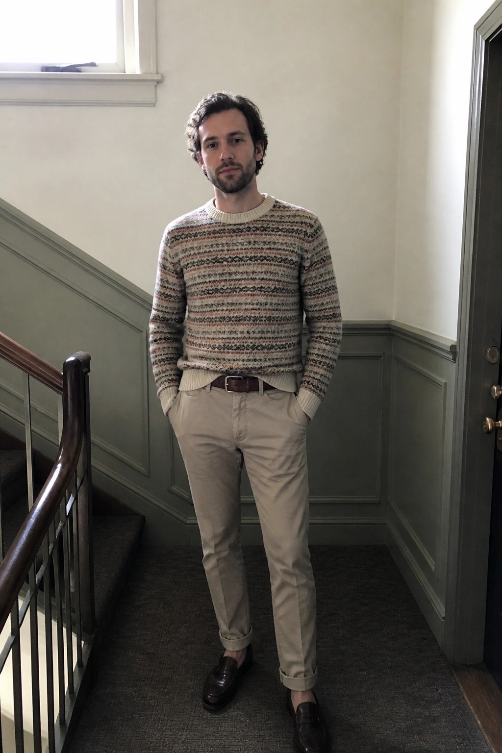 Man in patterned knit sweater and khaki pants stands in stairwell with hands in pockets