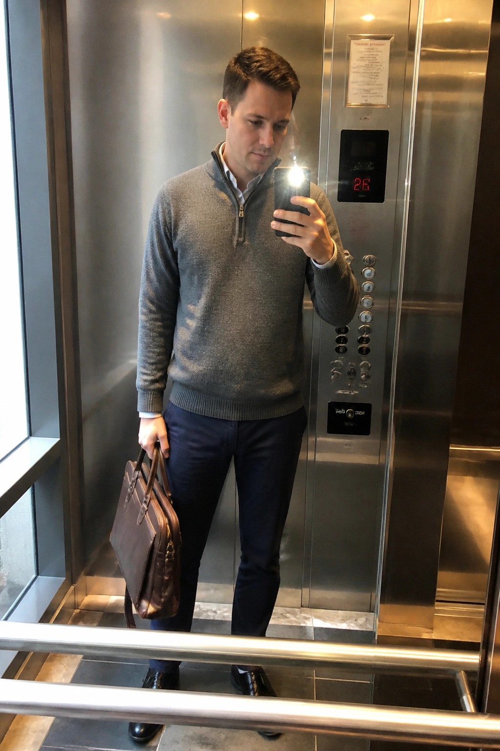 Man in gray quarter-zip sweater holding phone and brown briefcase in elevator