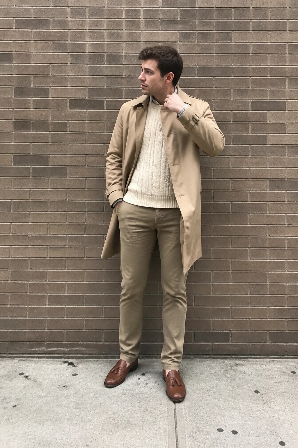 Man in beige trench coat and cream sweater stands against brick wall
