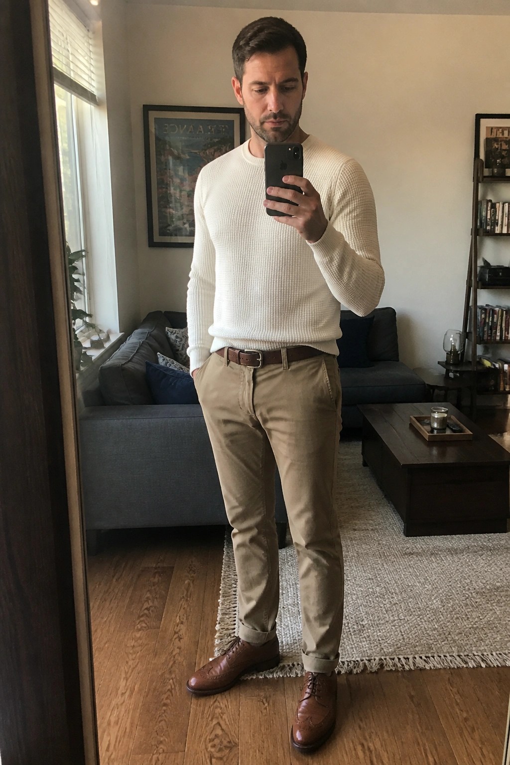 Man in white sweater and khaki pants taking mirror selfie in living room