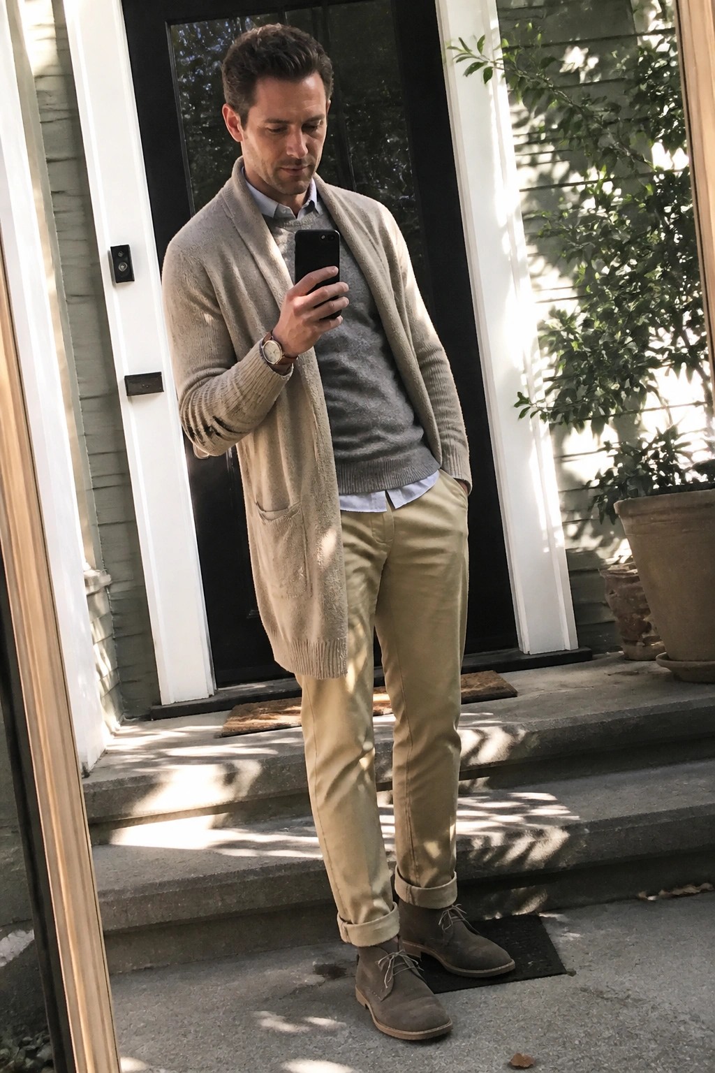 Man in beige cardigan layered over gray sweater and light blue shirt with khaki chinos and brown suede boots standing on steps while checking phone