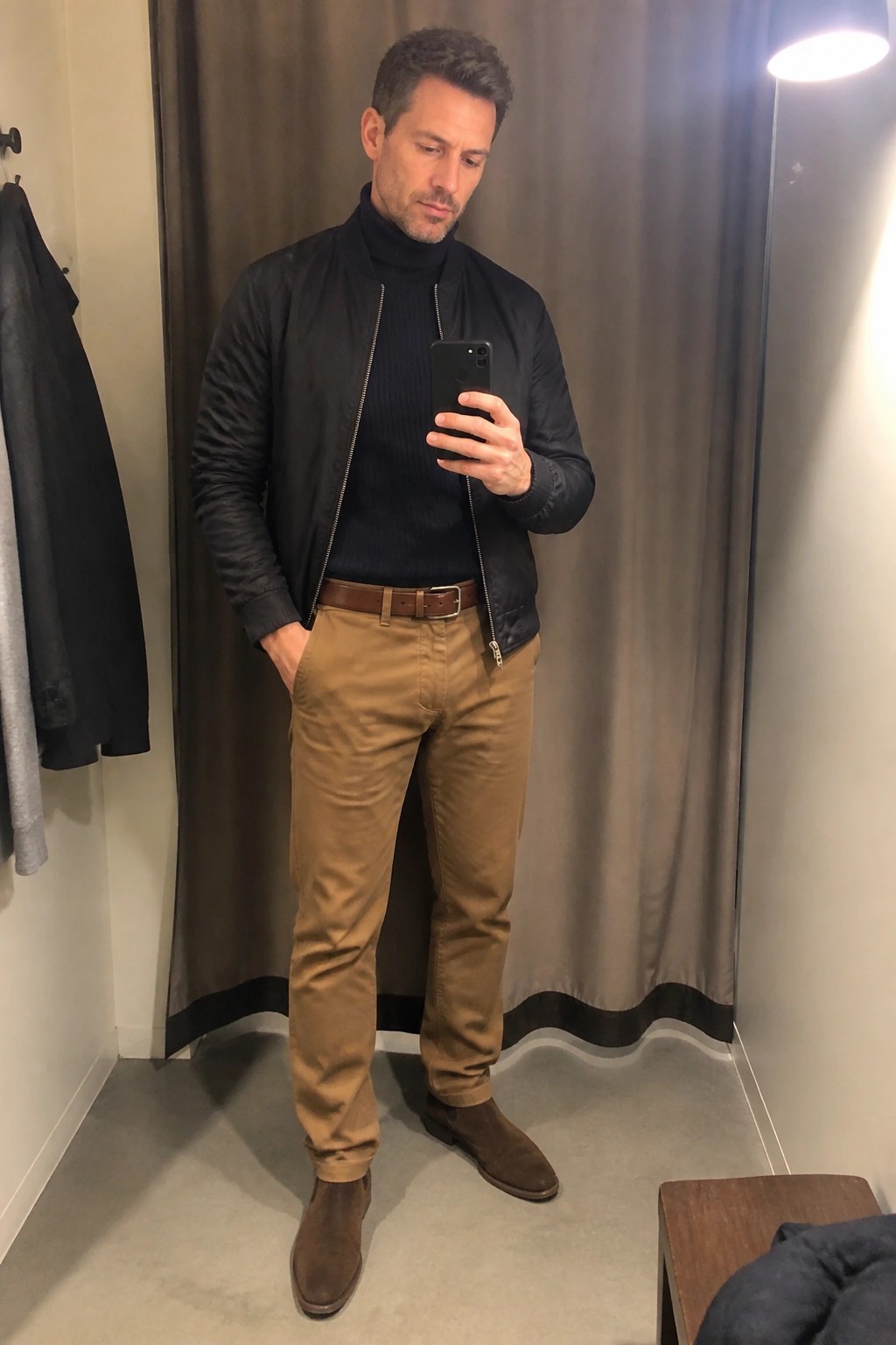 Man in black bomber jacket and tan pants taking mirror selfie in fitting room
