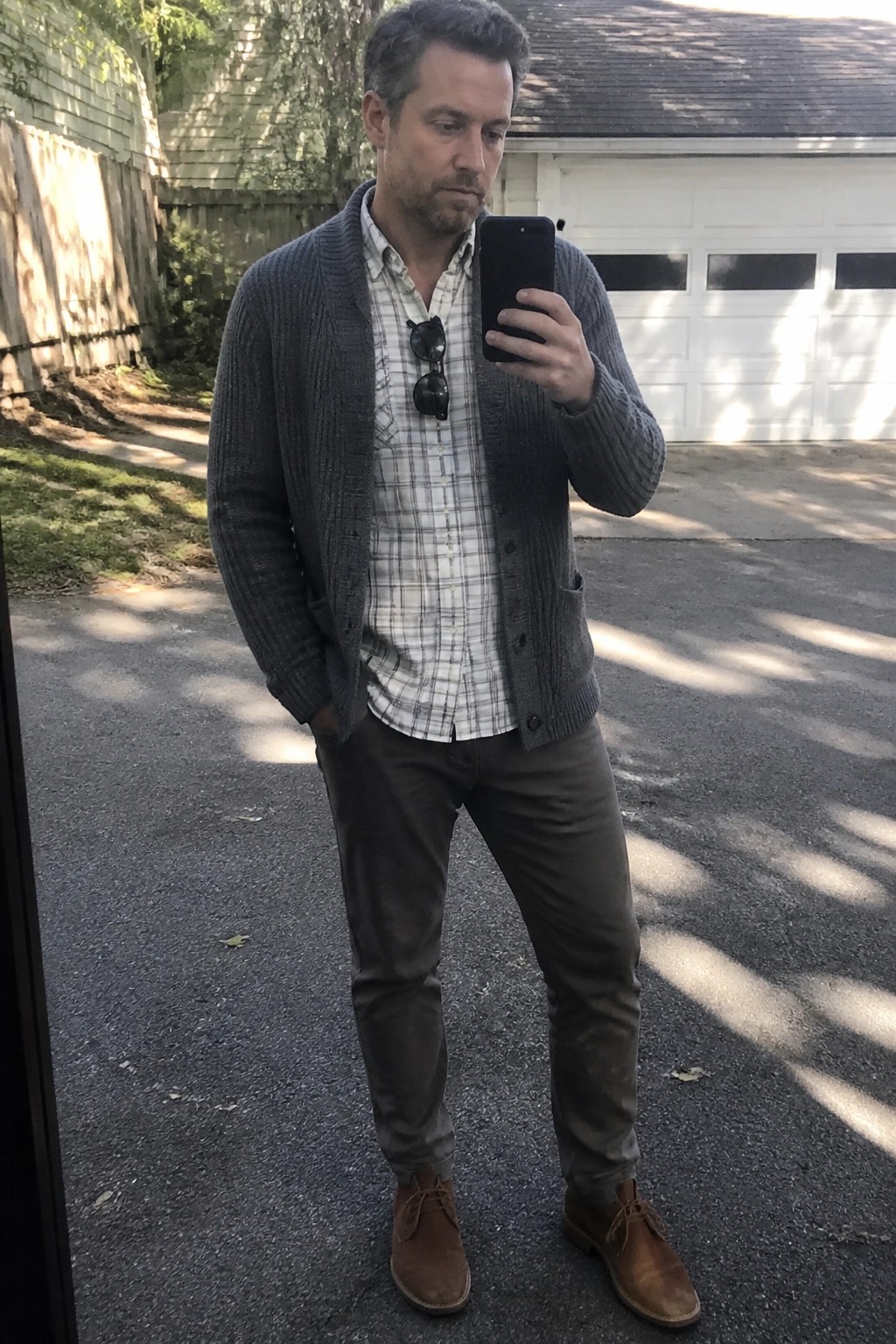 Man wearing an open gray cable knit cardigan over a white and gray plaid button up shirt tucked into olive chinos with brown suede chukka boots while holding a phone for a mirror selfie.