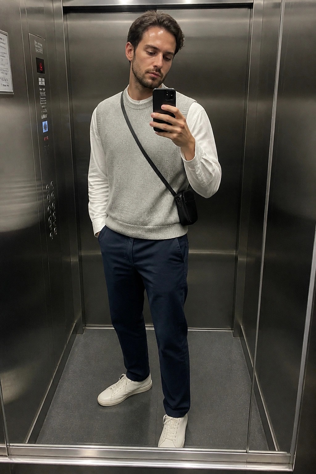 Man in gray sweater vest over white shirt taking mirror selfie in elevator