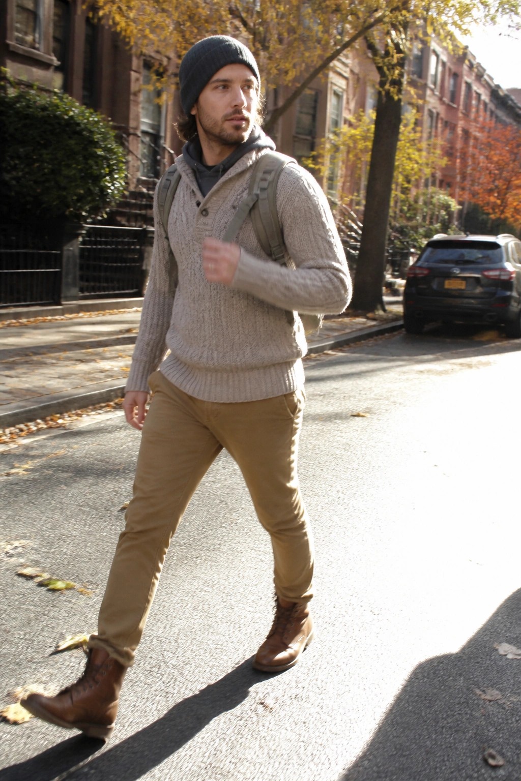 Man in gray knit sweater and beanie walks autumn city street with backpack.
