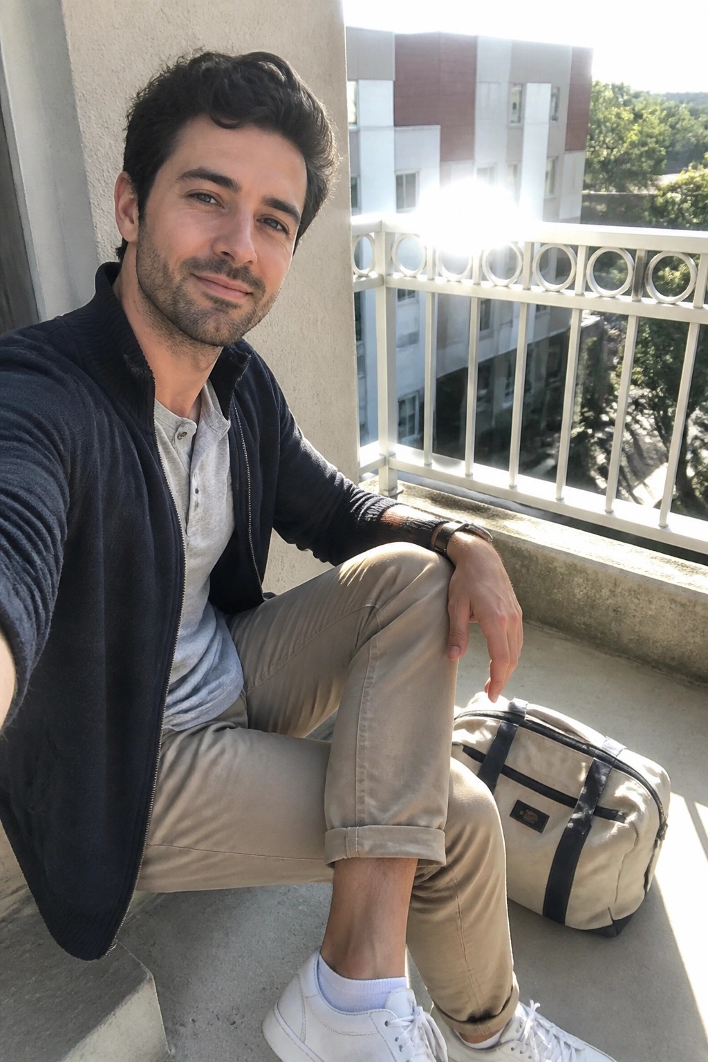 Man in navy sweater and beige pants sits on sunny balcony beside duffel bag