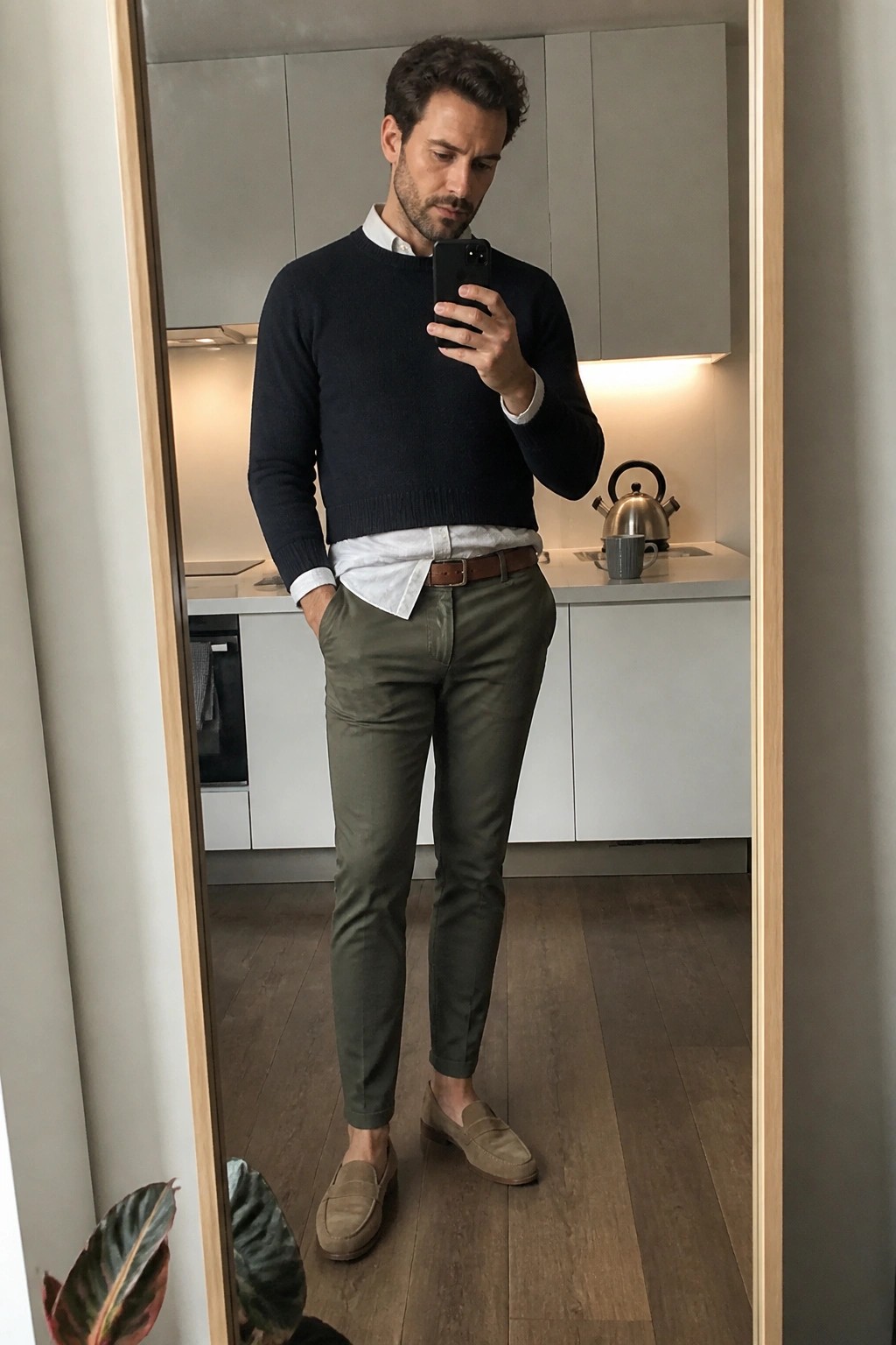 Man in navy sweater and olive pants taking mirror selfie in kitchen