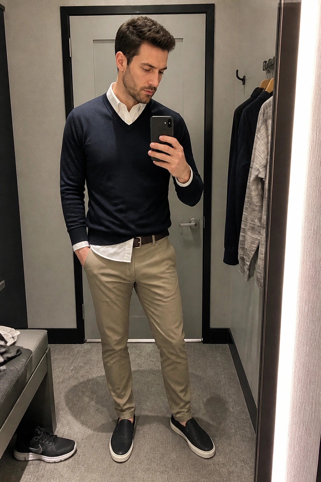 Man in navy sweater over white shirt and khakis taking mirror selfie in fitting room.