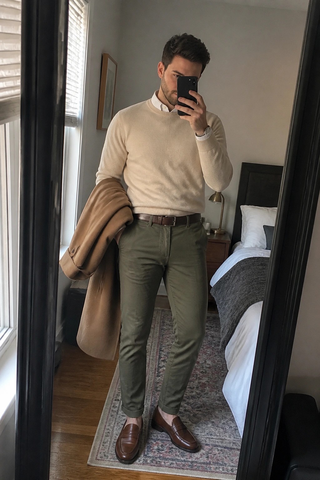 Man in beige sweater and olive pants taking mirror selfie while holding tan coat.