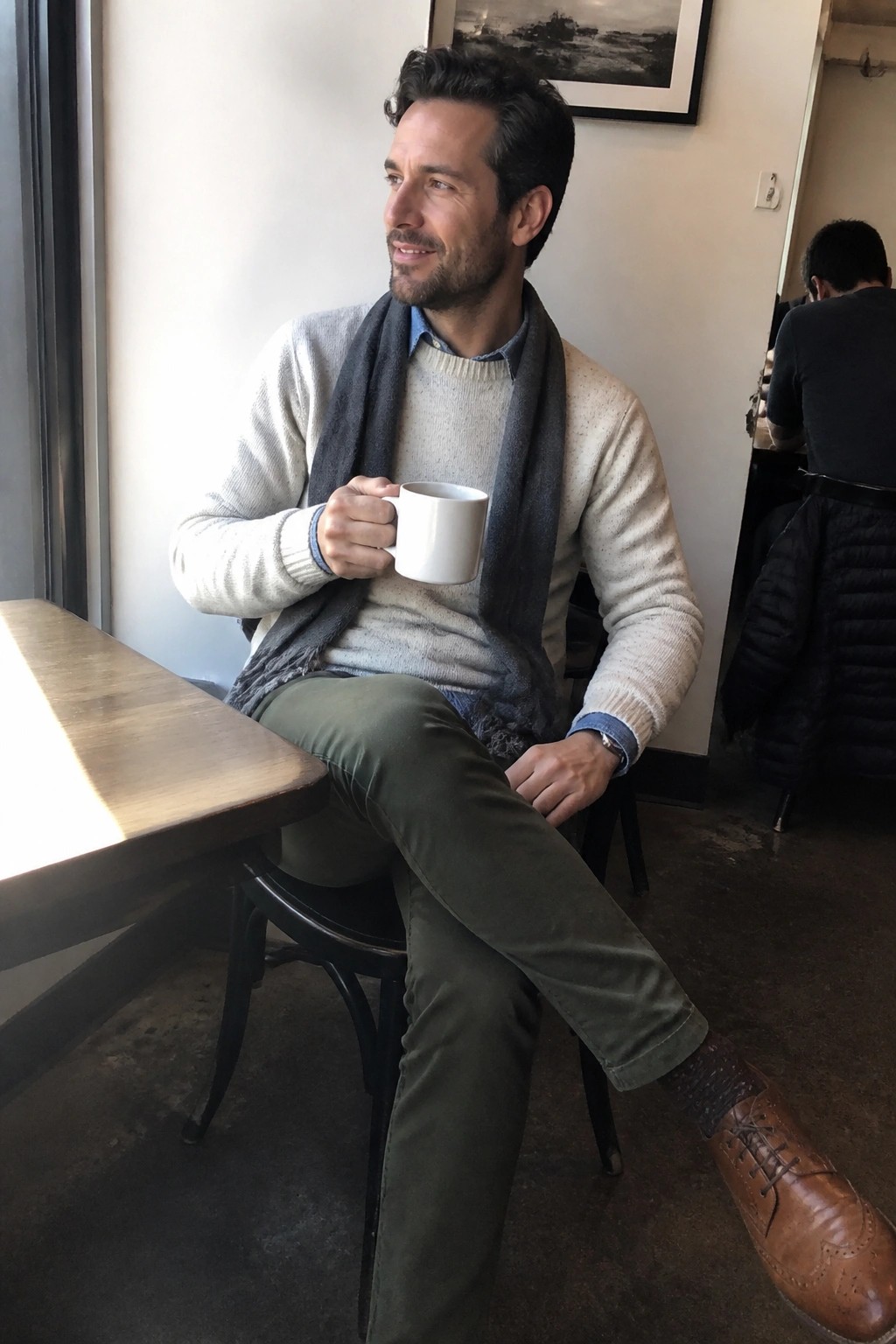 Man in cream sweater and gray scarf holding white mug at cafe table