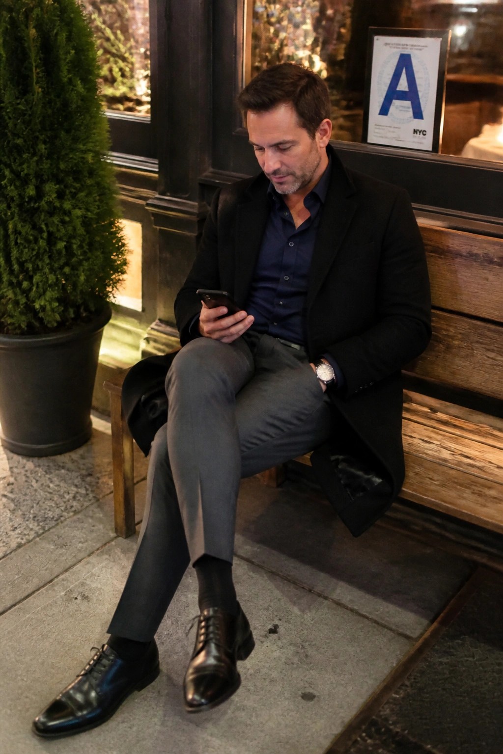 Man in black coat and gray pants views smartphone on wooden bench at night