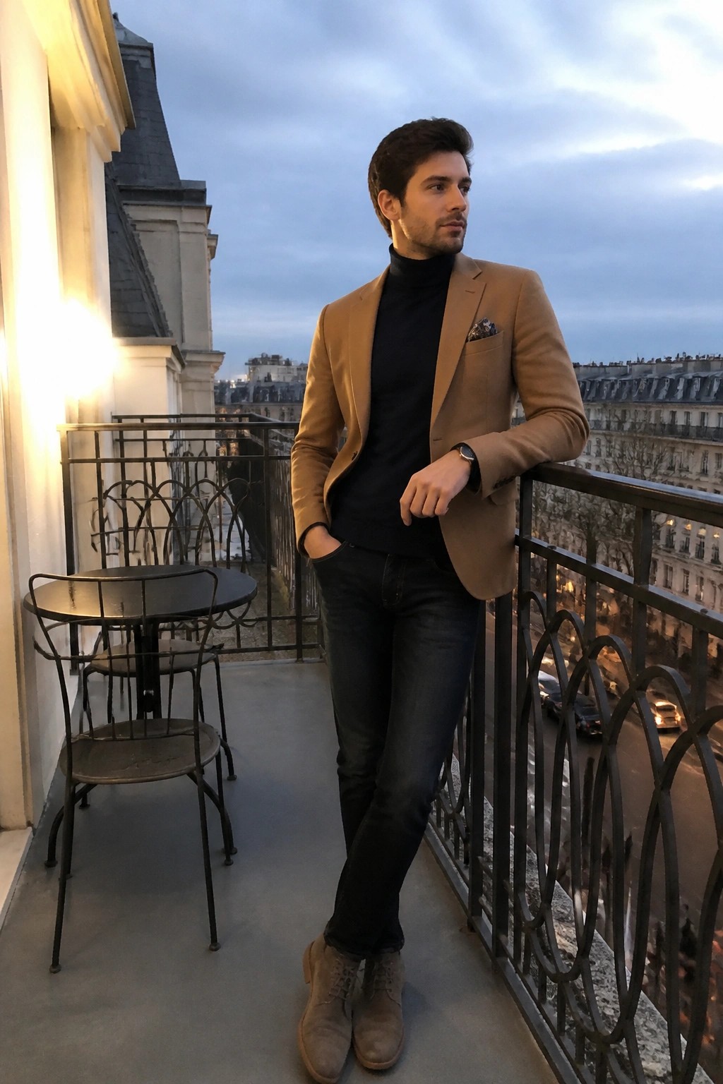 Man in tan blazer and black turtleneck on balcony overlooking city at dusk