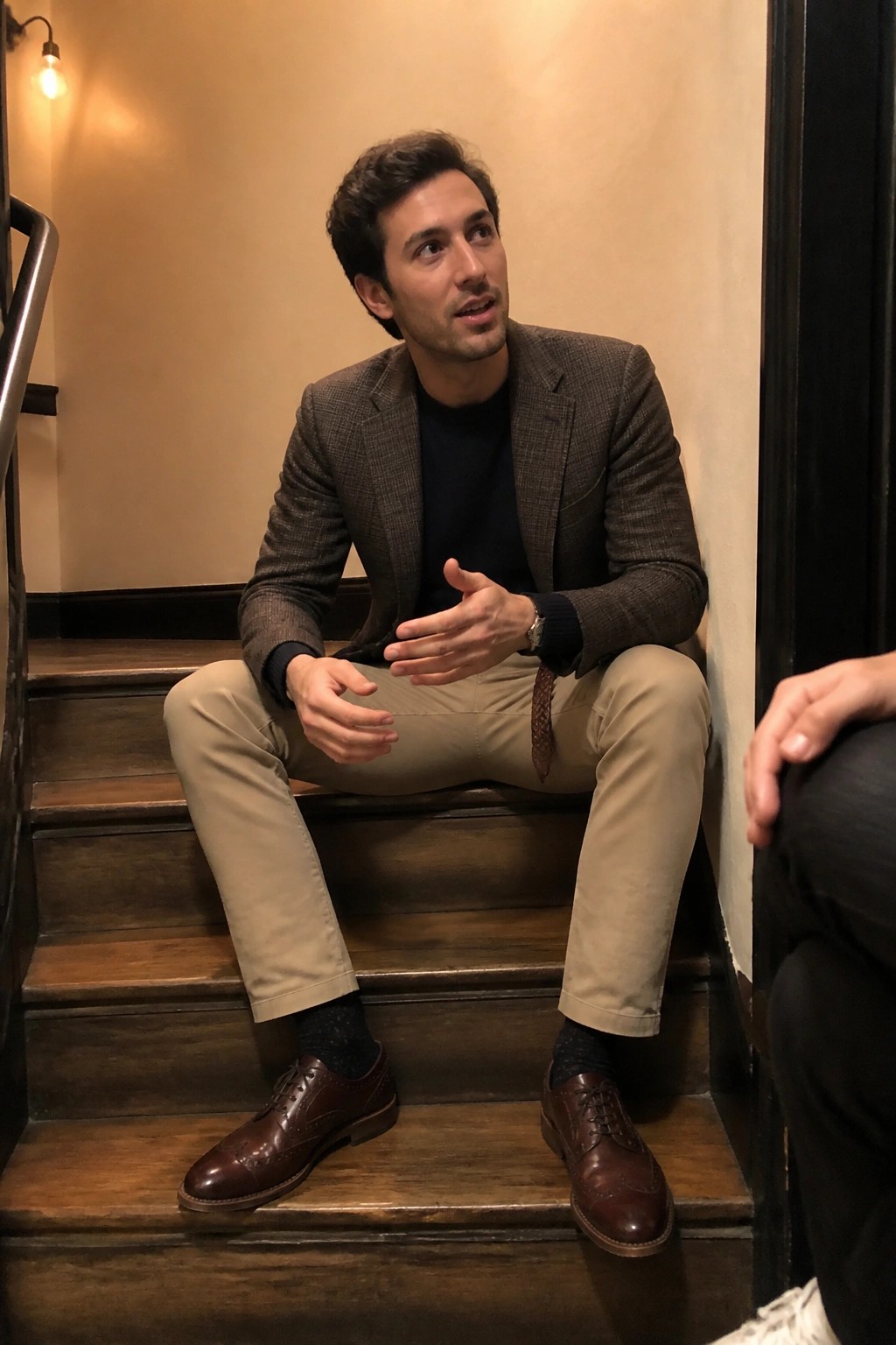 Man in plaid blazer and khakis gestures while seated on wooden stairs.
